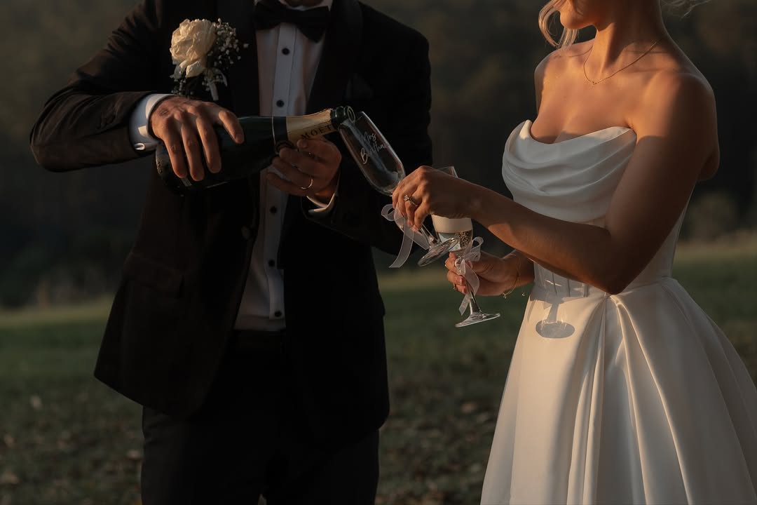 Wedding couple in black tie attire pouring champagne at sunset